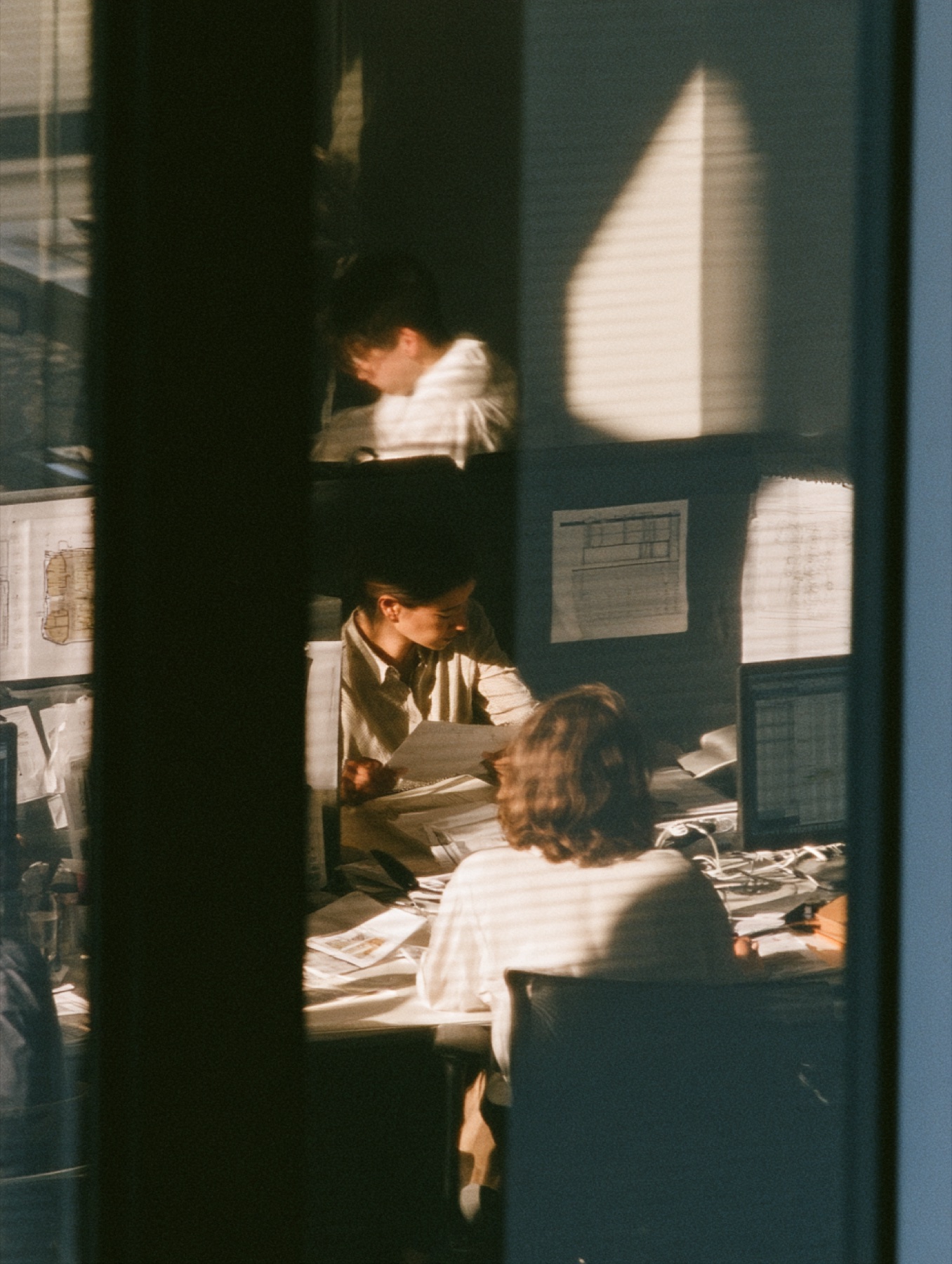 Colleagues reviewing drawings at a desk, seen through a window in late light