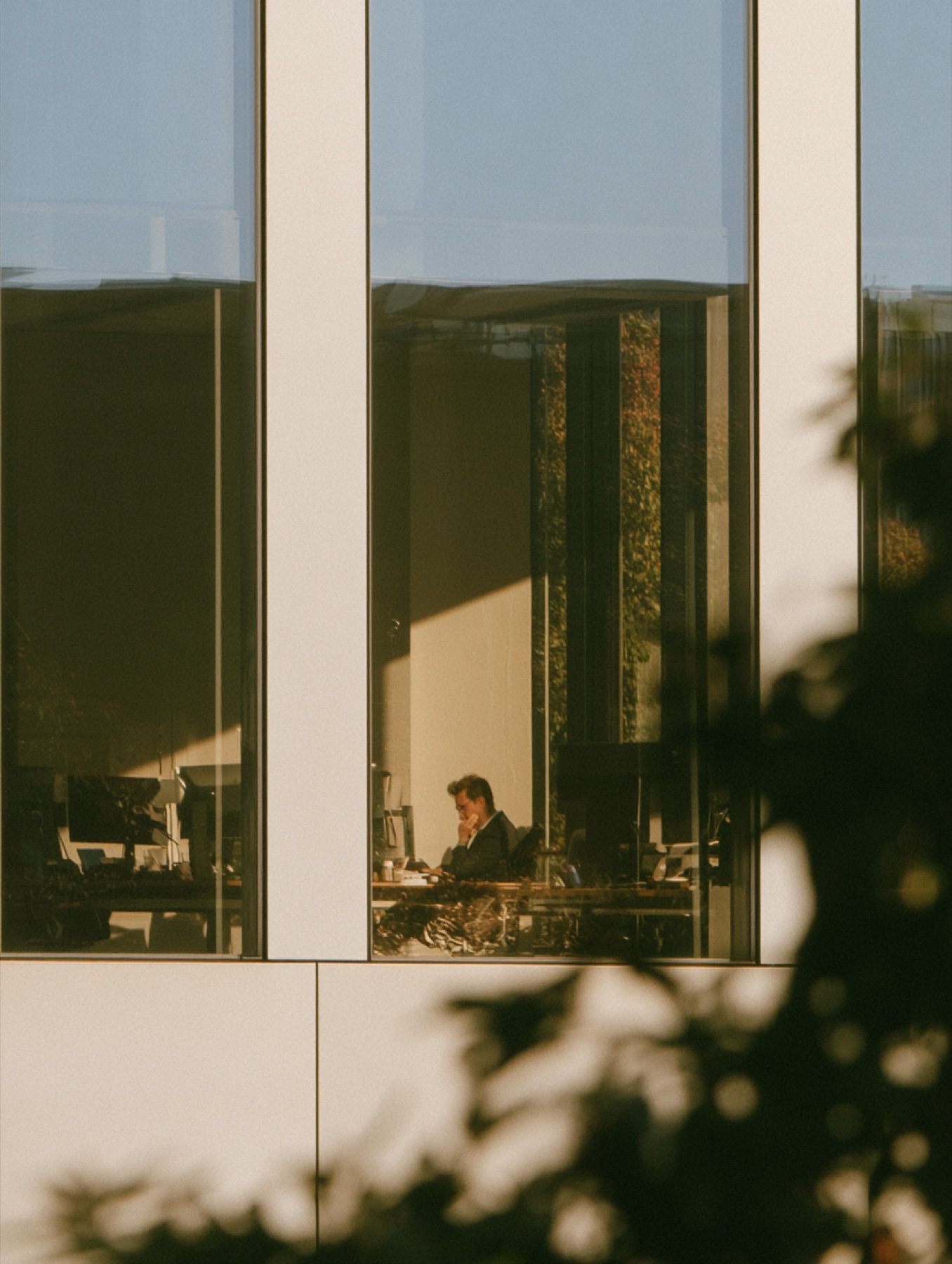 A person at a desk seen through a tall office window, leaves in the foreground
