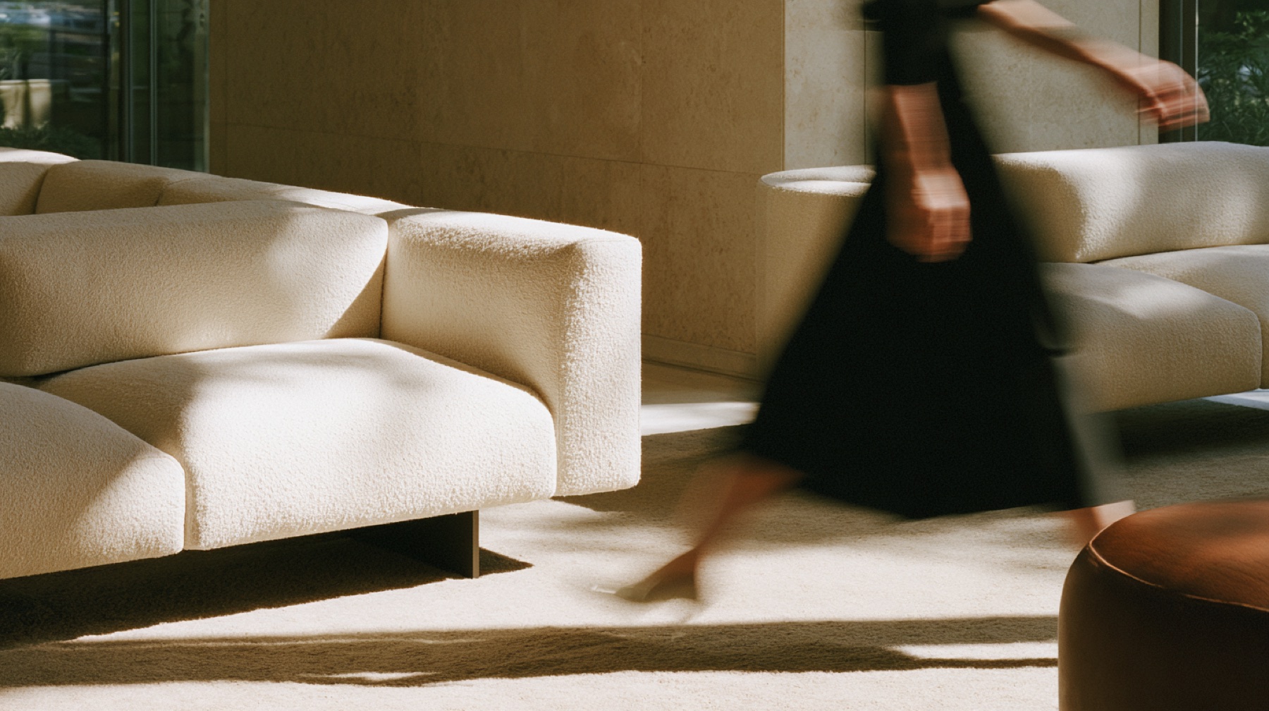Boucle sofas in a sunlit lobby, a figure in black passing in motion blur